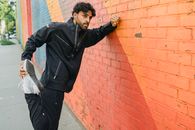 Man in Black Jacket and Black Pants Standing Beside Brown Brick Wall