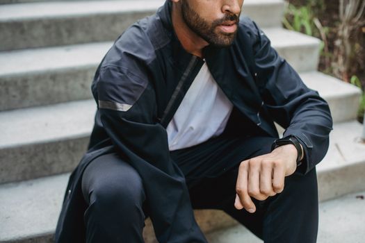 Close-up of a man sitting on steps wearing a stylish black sports jacket and digital watch.
