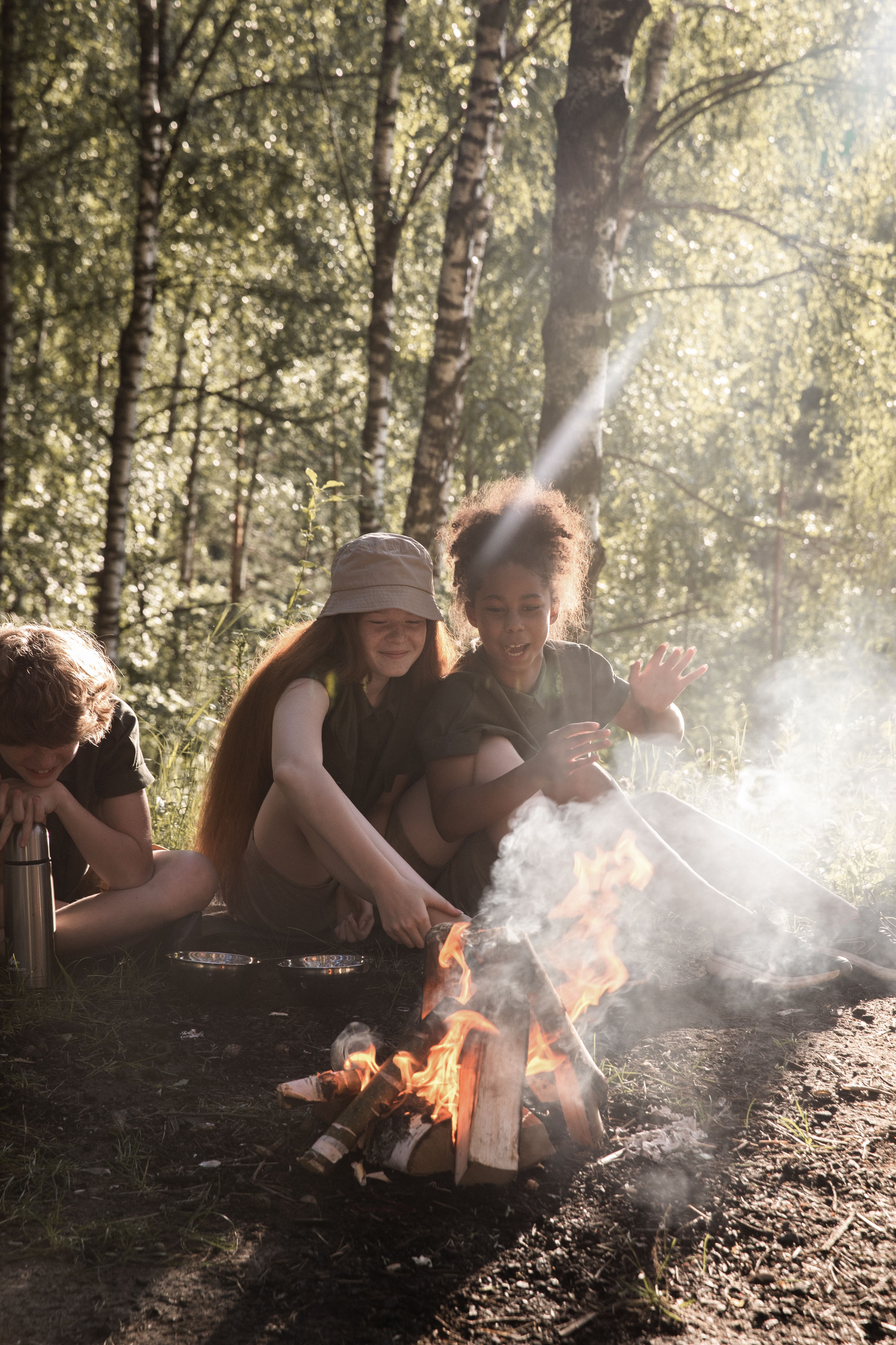 Children gather around a campfire in a sunny forest, enjoying nature together.