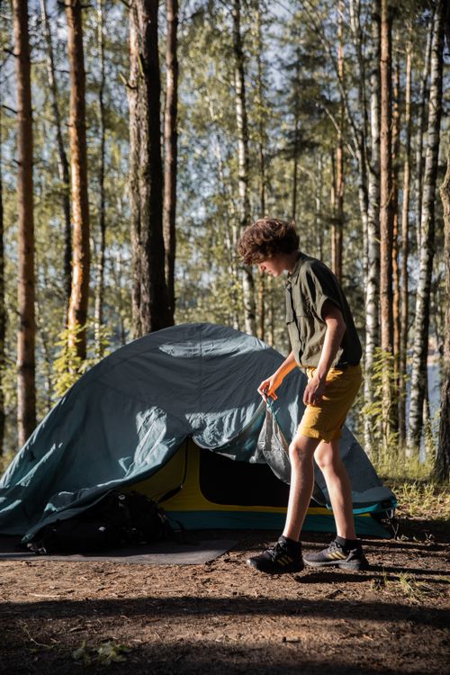 Free A young boy setting up a tent in a sunlit forest, ready for camping adventure. Stock Photo