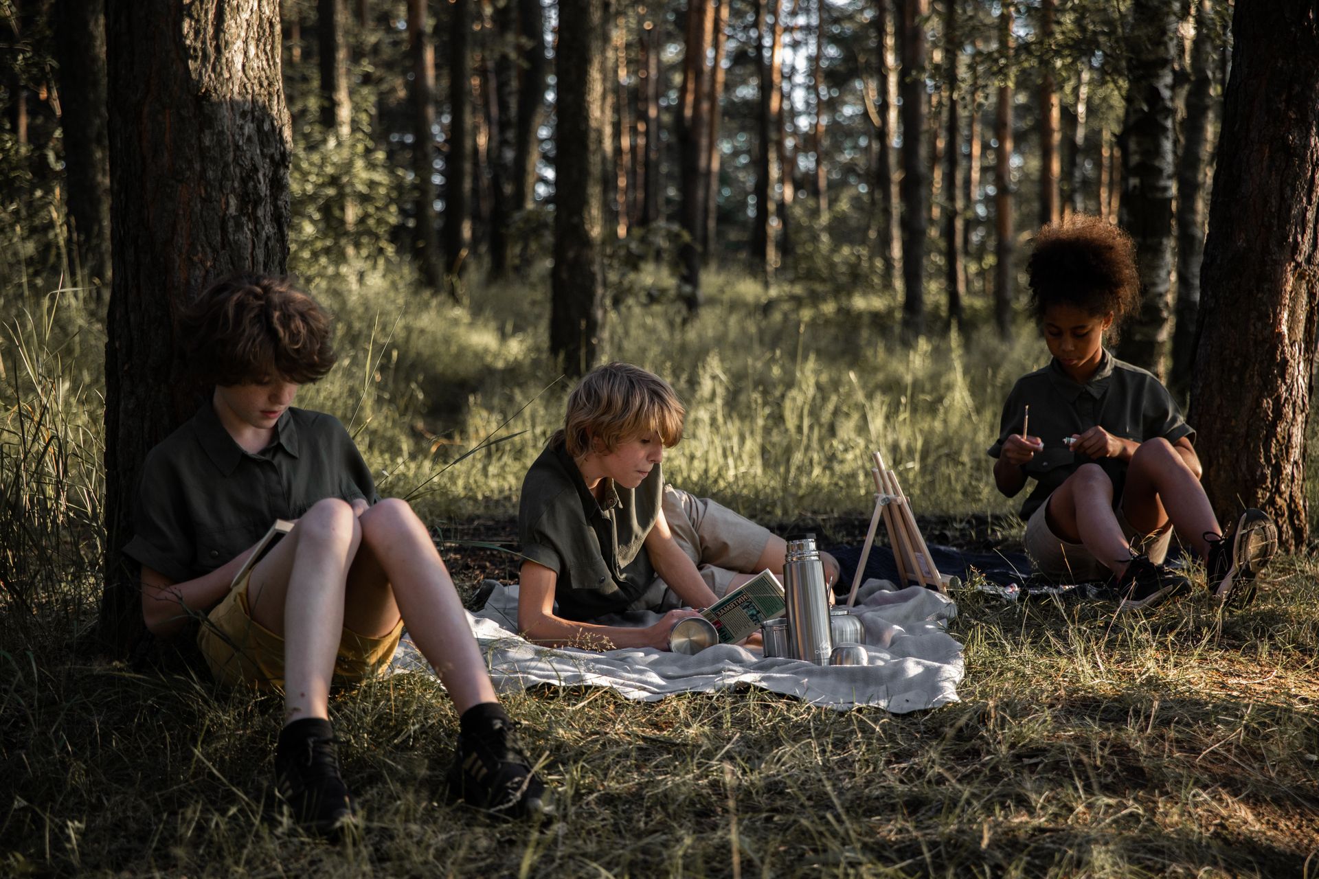 Three children engage in reading and crafting at a forest picnic. Ideal stock photo for education and adventure themes.