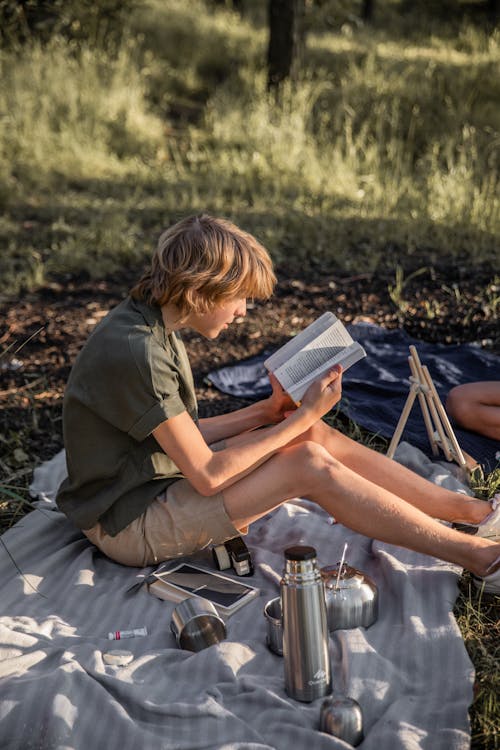 A Boy Sitting on a Picnic Blanket while Reading a Book