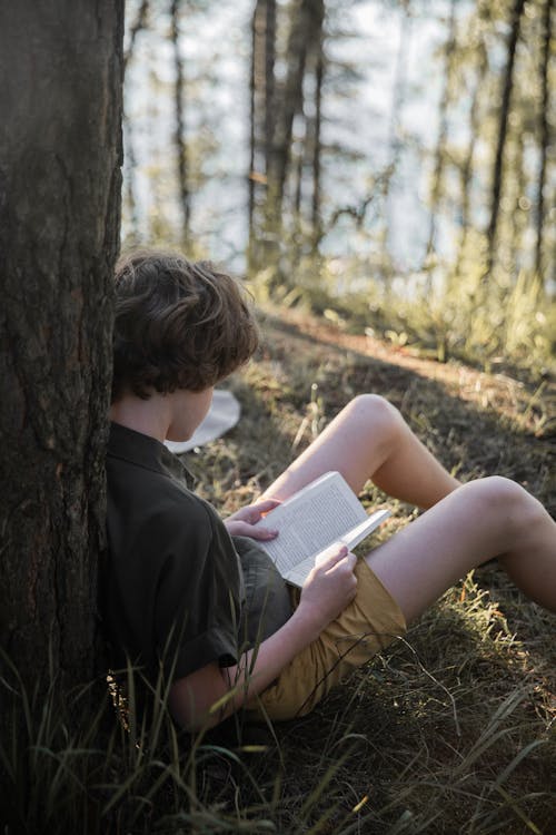 Boy in Khaki T-Shirt Reading Book While Sitting on the Ground Beside Tree Trunk