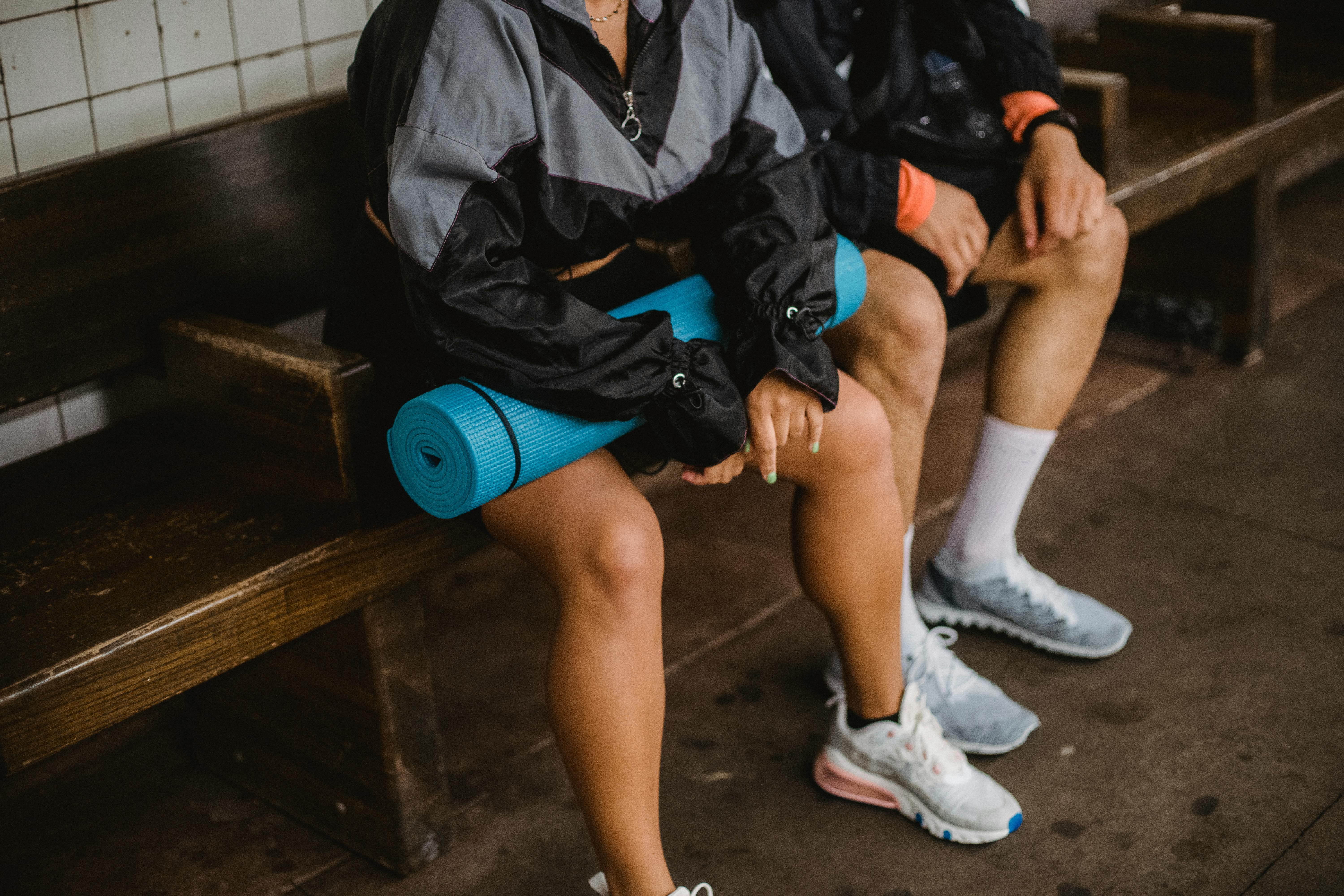 Two people in sportswear sitting on a bench, holding a yoga mat, showcasing athletic lifestyle.