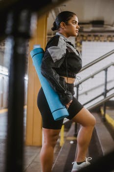 Young woman in athletic clothing carrying a blue yoga mat while ascending subway stairs.