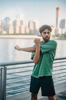 A man in a green shirt stretches by the waterfront with a city skyline in the background.