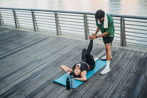 Free Man assisting woman in stretching exercises on boardwalk next to river, emphasizing fitness and teamwork. Stock Photo