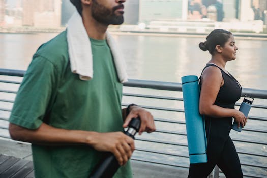 Two friends walking outdoors by the water, carrying yoga gear and bottles, embracing a healthy lifestyle.