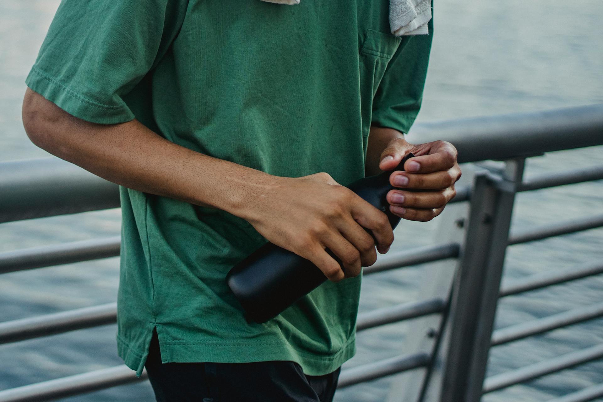Close-up of a man holding a black water bottle by a waterfront rail in casual attire.