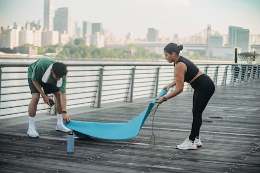 A man and woman set up a yoga mat on an urban boardwalk with a city skyline backdrop.