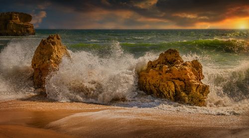Free Powerful ocean waves crash against rugged rocks under a vibrant sunset sky, creating a dramatic seascape. Stock Photo