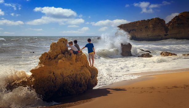 Three children climb rocks as waves crash on a sunny beach day, enjoying nature.