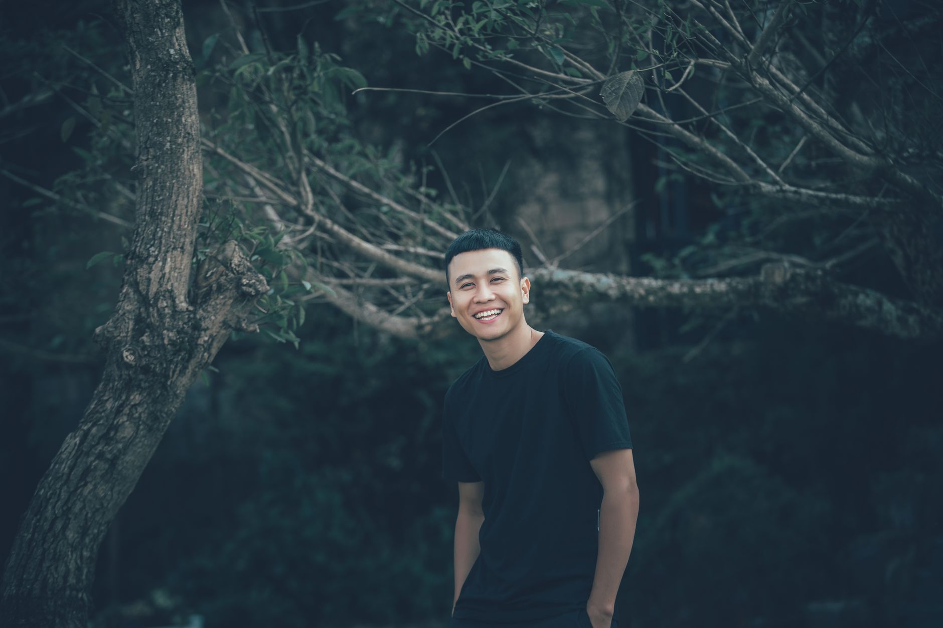 A cheerful man poses outdoors in a natural setting, wearing a black t-shirt and smiling warmly.