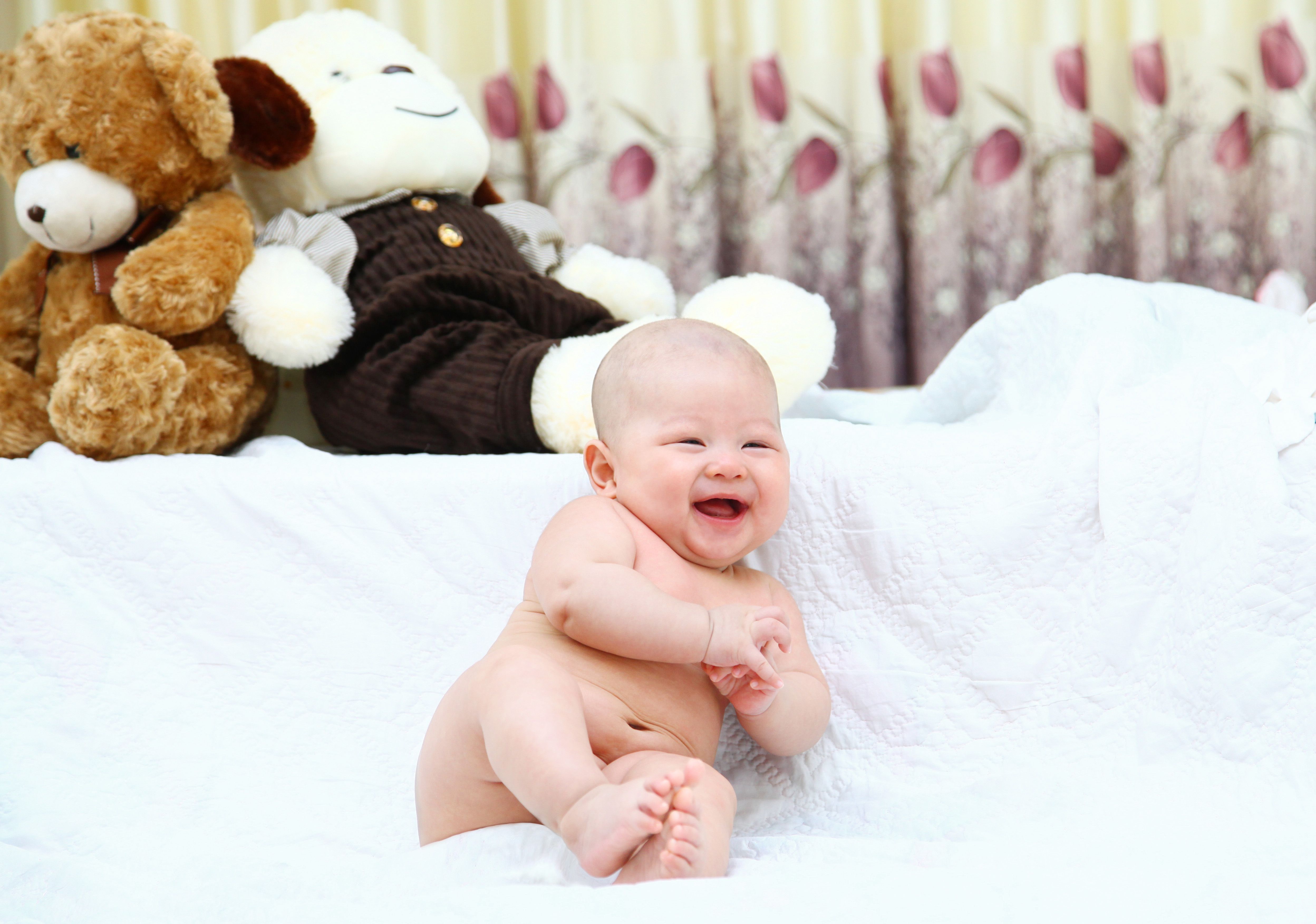 Cute smiling baby sitting on a white blanket with teddy bears in the background.