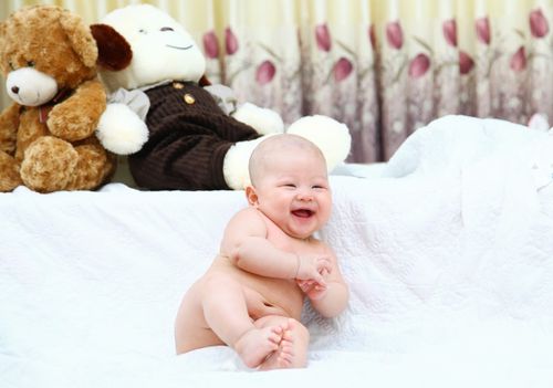 Free Cute smiling baby sitting on a white blanket with teddy bears in the background. Stock Photo
