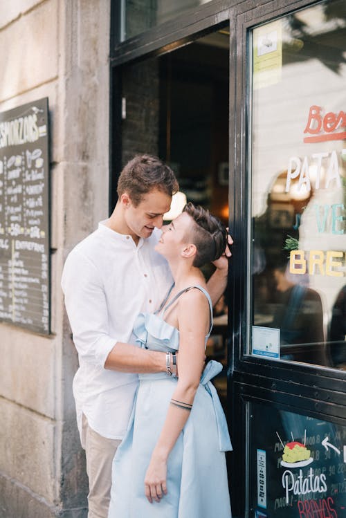 Free Man in White Dress Shirt Hugging a Woman in Blue Sleeveless Dress Outside a Bakehouse Stock Photo