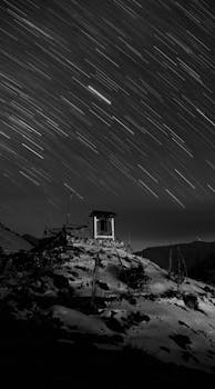 Black and white photo of star trails over a snow-covered mountain landscape at night.
