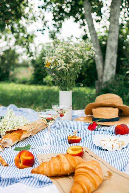 Brown Straw Hats Beside White Flowers and Fruits with Two Croissants on Picnic Blanket