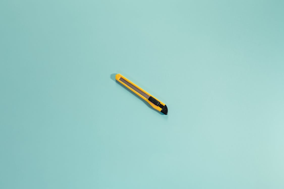 From above of yellow stationery knife with sharp blades placed in center of bright empty background in daylight in modern studio