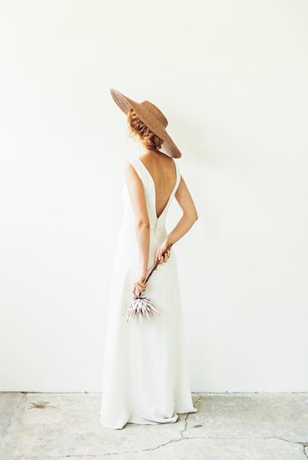 A stylish woman in a backless white dress and sun hat holds dried flowers, standing against a minimalist backdrop.