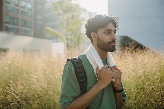 A young man stands outdoors in sunlight, wearing casual sports clothing and carrying a backpack.