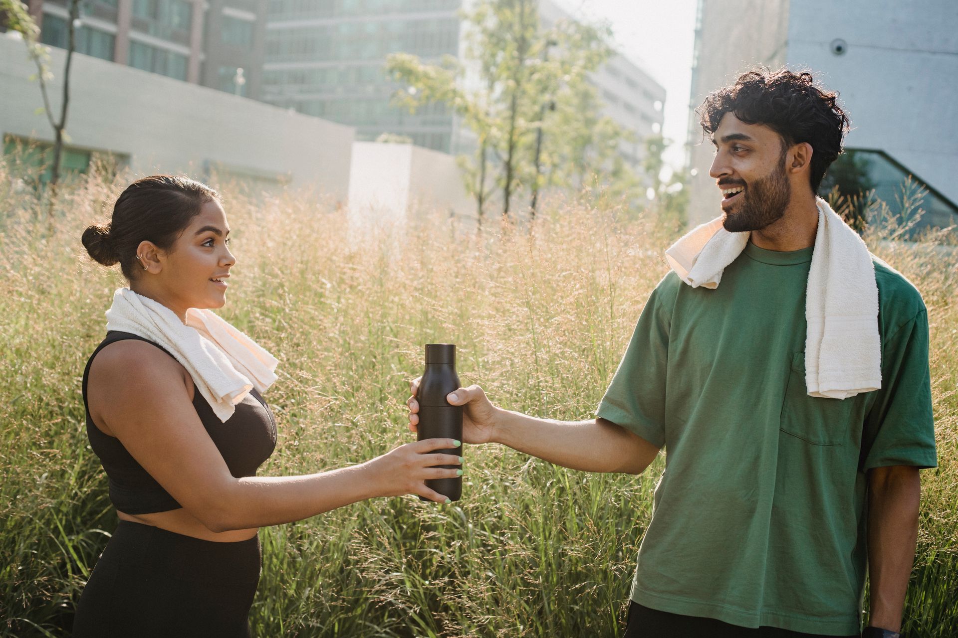 Two friends in sportswear share a water bottle in a city park on a sunny day.