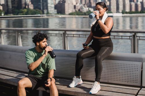 Free A young couple takes a break by the waterfront to hydrate after exercising outdoors in a sunny city setting. Stock Photo
