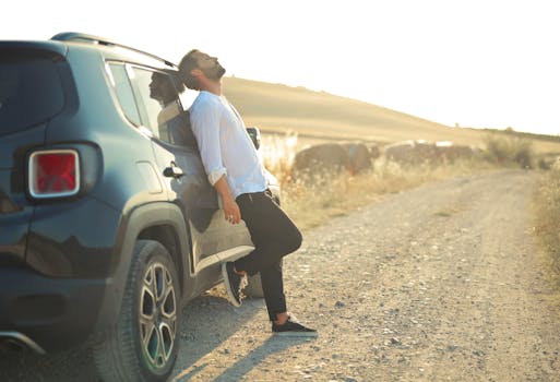 A man leans back on his SUV parked on a dirt road during a serene sunset, embodying relaxation and travel.