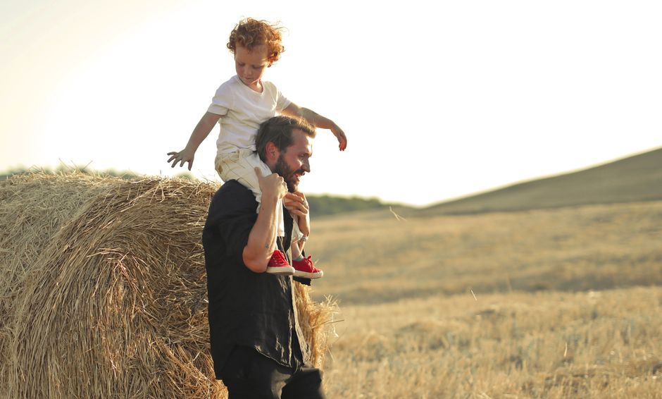 Padre llevando a su hijo a caballito a través de un soleado campo rural, disfrutando de un día de verano juntos.