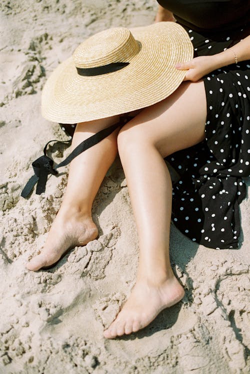 Woman in Black and White Polka Dot Skirt with Brown Sun Hat Sitting on Sand 