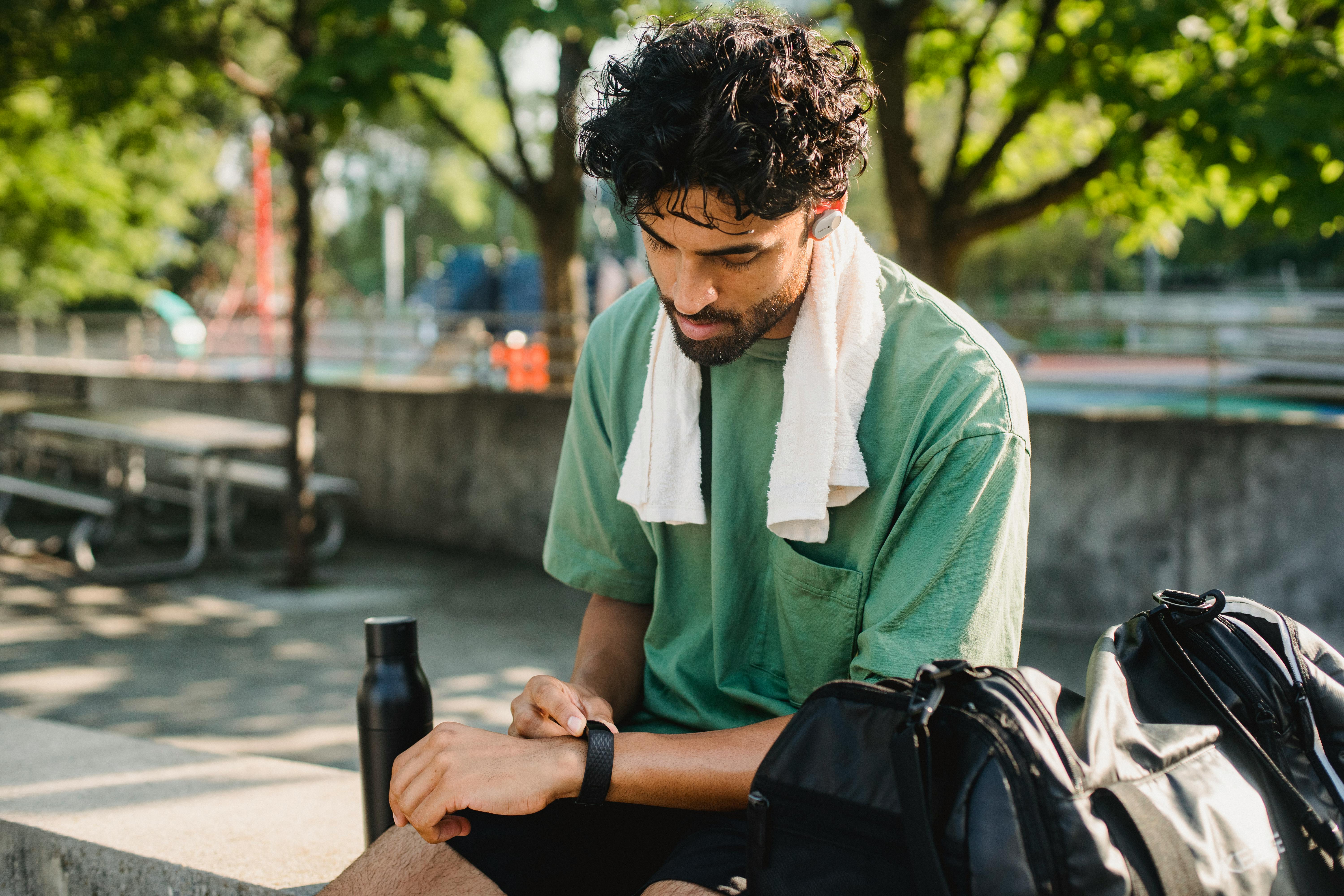 Man resting on a park bench after workout with green shirt, towel, and water bottle.
