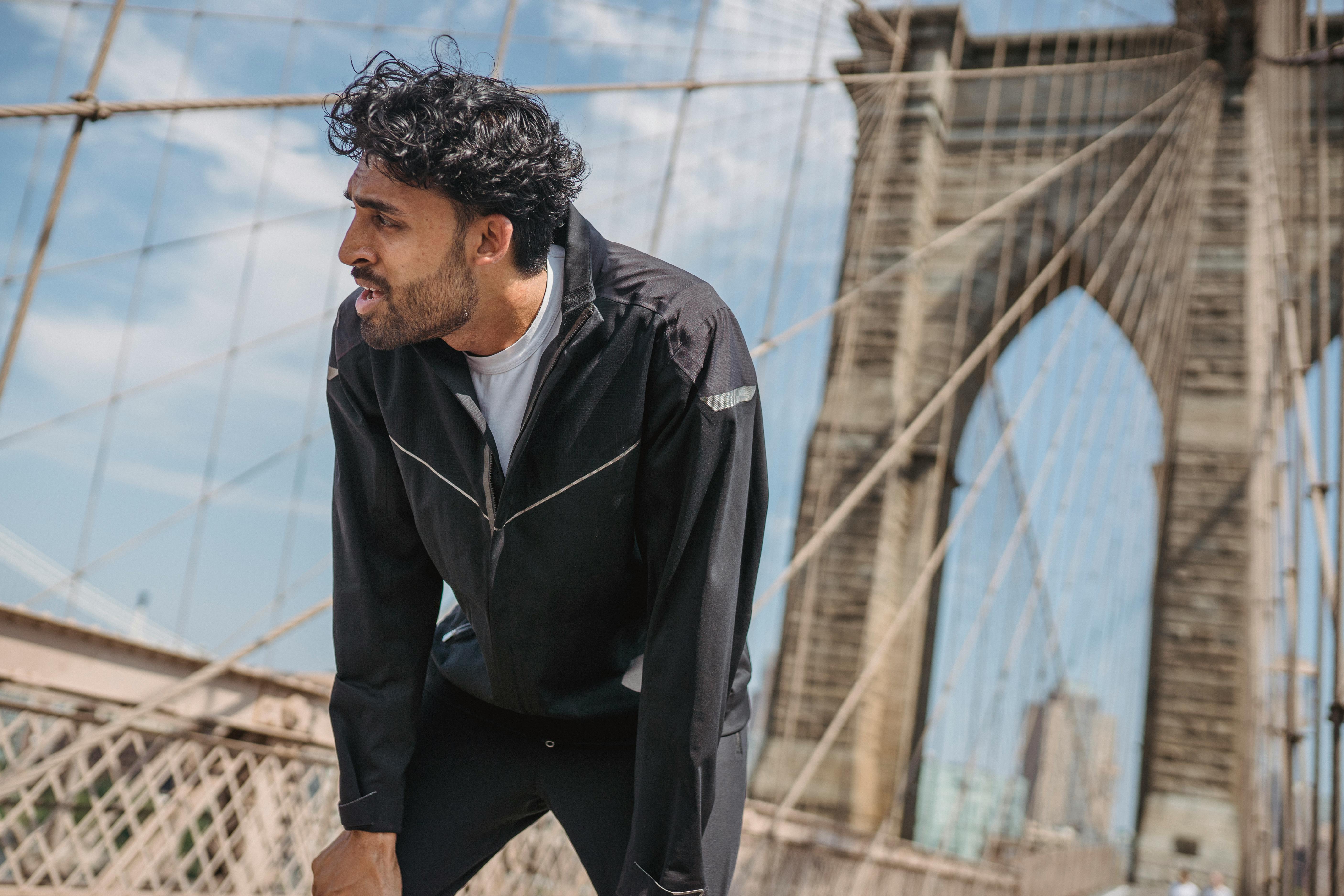 一名穿着黑色夹克的男子站在布鲁克林大桥上，背景是模糊的城市景观