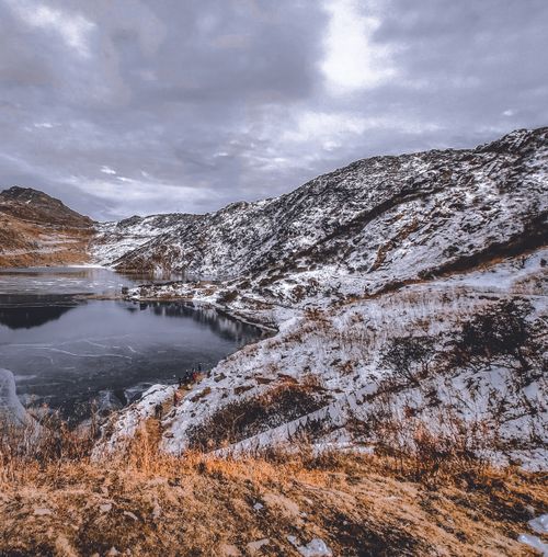 Free A tranquil winter landscape featuring a serene lake surrounded by snow-covered mountains under a cloudy sky. Stock Photo