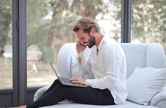 Caucasian man in white shirt smiling while working with laptop and phone in a cozy indoor setting.
