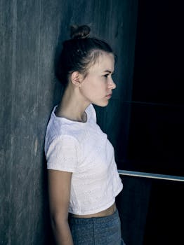 Side view of a young woman in casual attire leaning against a wall, lost in thought.