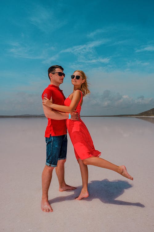 Full body of young barefoot travelers in casual clothing cuddling on wet sand under blue sky covered with clouds