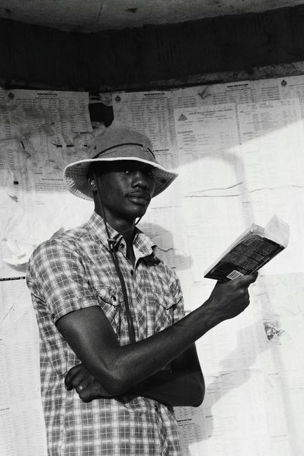 Black and white of serious African American male in hat looking at camera while standing near board with papers with book in hand