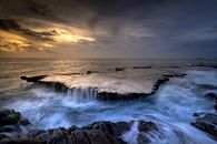 Majestic scenery of rough rocky coast and stormy foamy sea under dark cloudy sky with sun beam at sundown