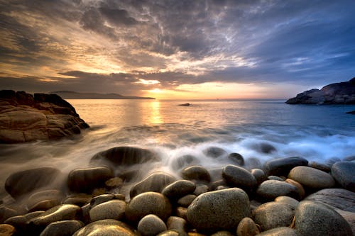 Picturesque scenery of cloudy sky and big heavy stones and rocks washing by foamy waves  of ocean at sundown