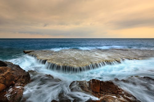 Picturesque scenery of endless ocean with foamy waves washing rocky coast against cloudy sunset sky