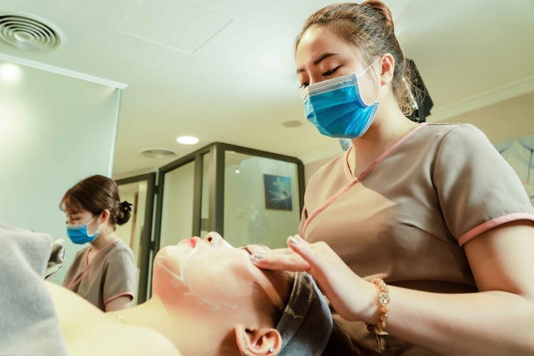 A Woman Wearing Face Mask While Putting A Cream On Her Client's Face