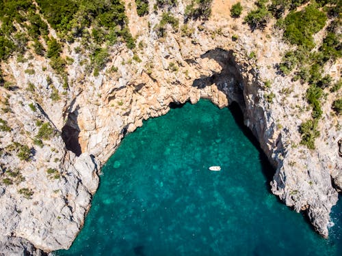 Breathtaking drone view of white yacht sailing on emerald sea surrounded by rough rocky cliff with green trees on top on sunny day