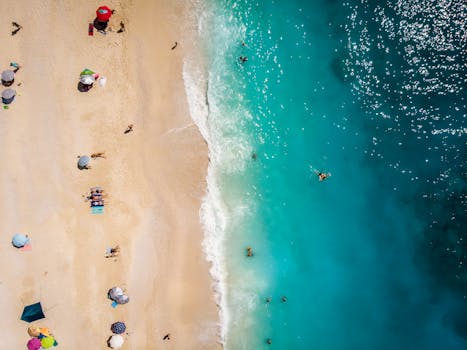 Drone aerial shot capturing sunbathers and turquoise waves on a sandy beach.