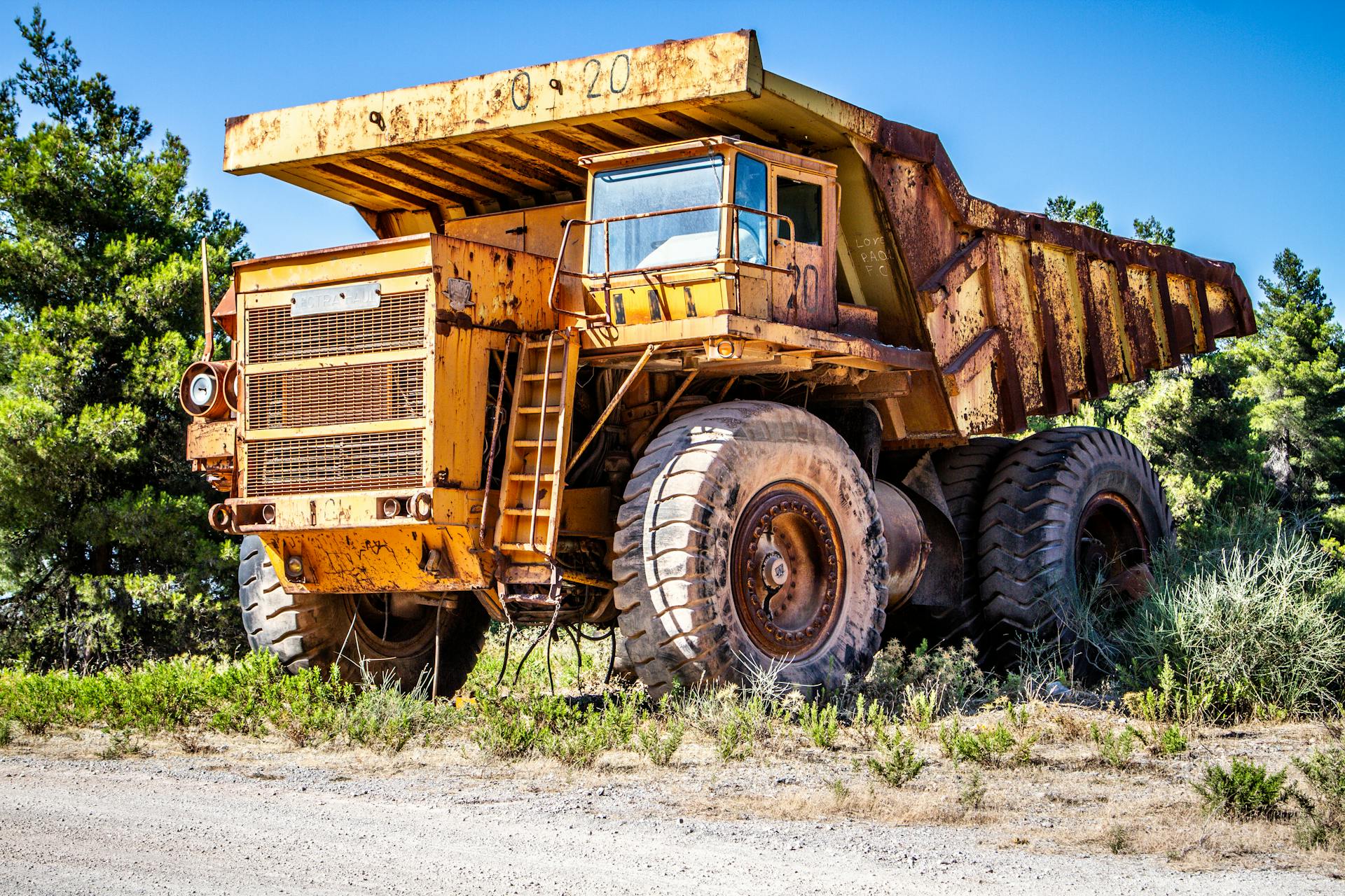 Old rusty dump truck parked in a sunny countryside area with clear sky.