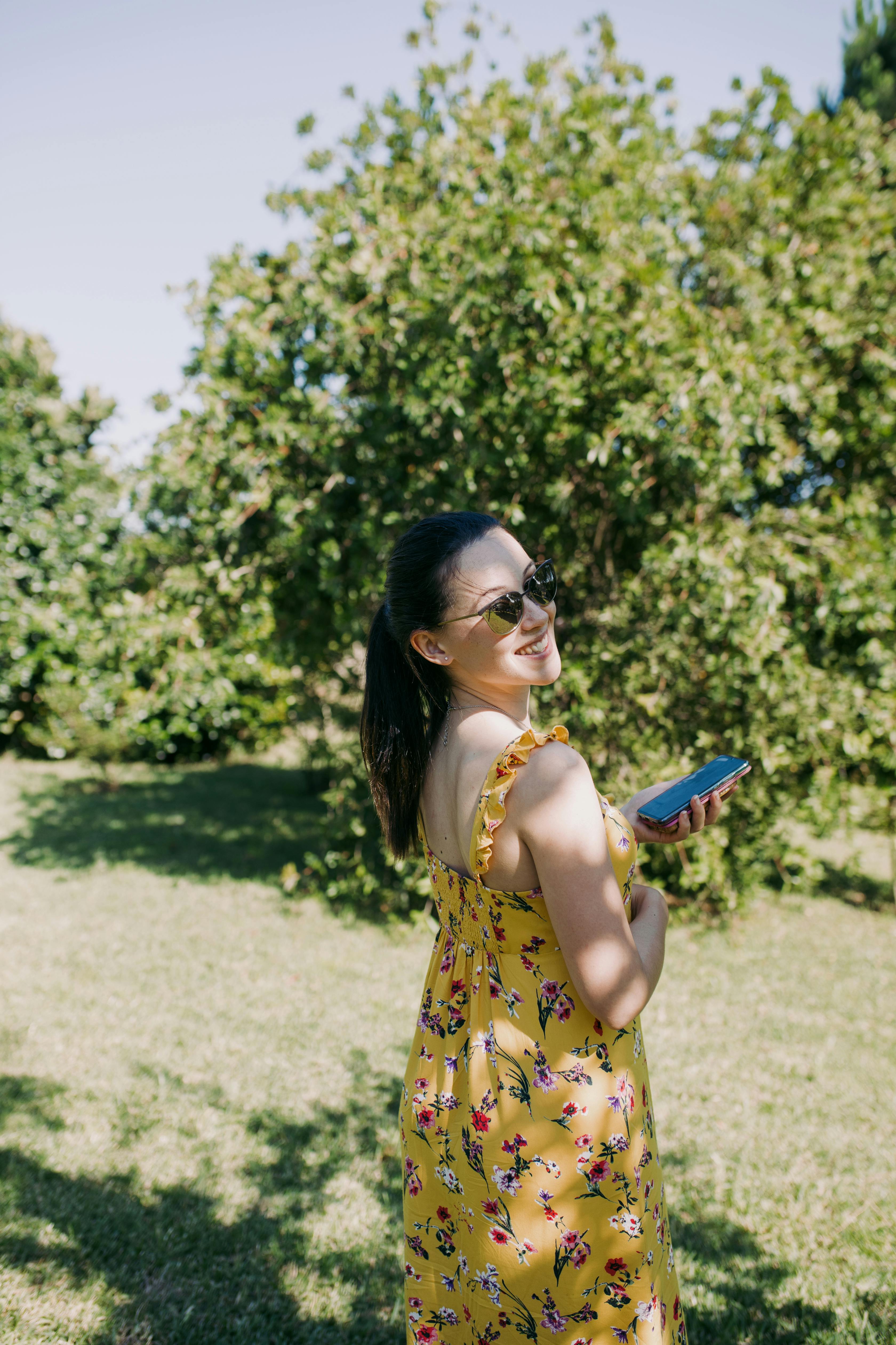 Woman in floral yellow dress holding a smartphone and smiling outdoors.