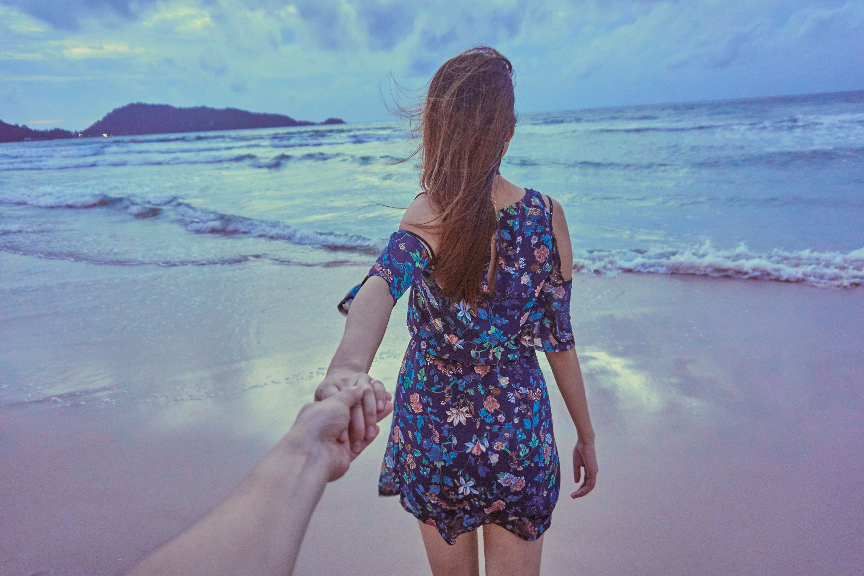 A woman in a floral dress leading a partner on a Phuket beach at dusk, holding hands, with waves in the background.