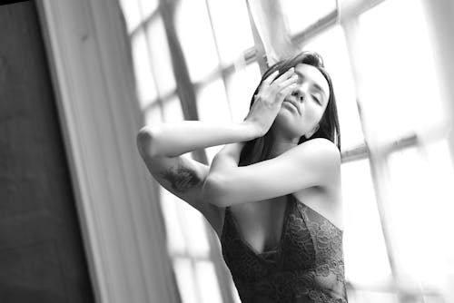 Black and white sensual attractive female touching face gently while standing with eyes closed near window in studio