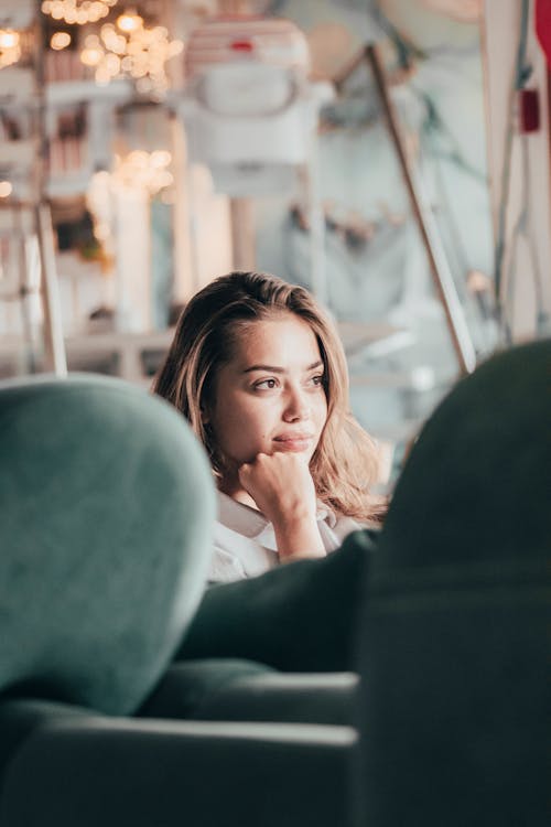 Charming young female sitting on comfy couch in modern interior and resting head on hand while looking away in dreams