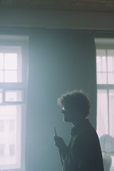 Silhouetted man standing by window, holding an object, creates a moody indoor scene.