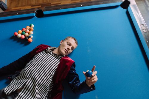 Free Adult man in a striped shirt lying on a blue billiard table holding a cue ball. Stock Photo